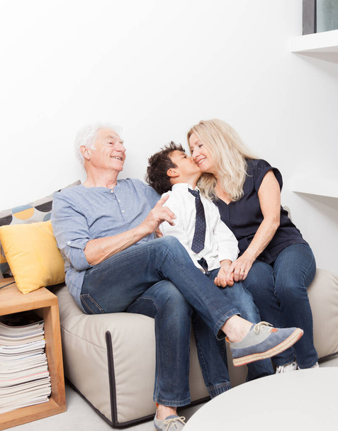 Мальчик целует её бабушку. Пенсионеры с внуком на диване в гостиной. Концепция семьи, разделения и радости
 - Фото, изображение