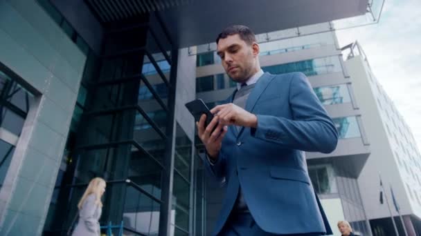 Caucasian Businessman in a Suit is Using a Smartphone on a Street in Downtown. Other Office People Walk Past. He Smiles and Looks Successful. He's Browsing the Web on his Device. - Filmati, video