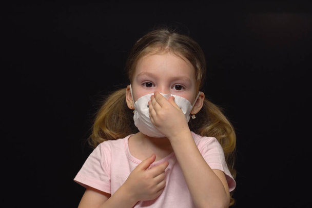 Концепция коронавируса и загрязнения воздуха. Маленькая девочка в маске для защиты. Коронавирус Ухана и симптомы эпидемии вируса. Регулирует маску рукой. Изолированный на черном фоне
. - Фото, изображение