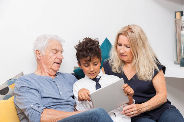 Authentiek moment wanneer jongen met haar grootmoeder en zijn grootvader spelen met een digitale tablet op de bank. Lachende familie met tablet thuis - Foto, afbeelding