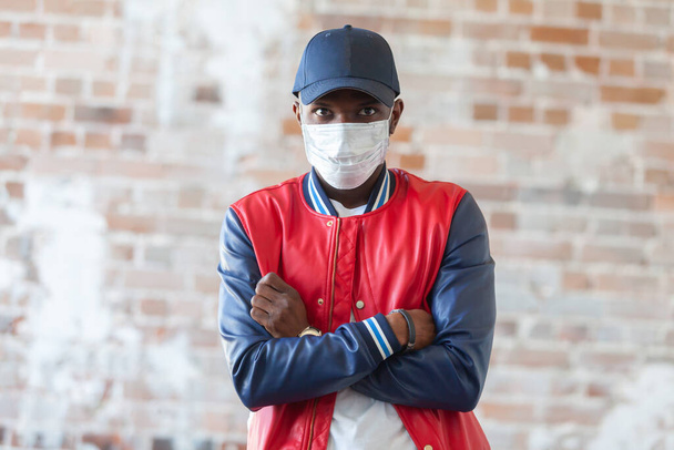 Handsome african man wearing medical mask is shocked with coronavirus 2019 danger on background of ruined red brick wall - 写真・画像