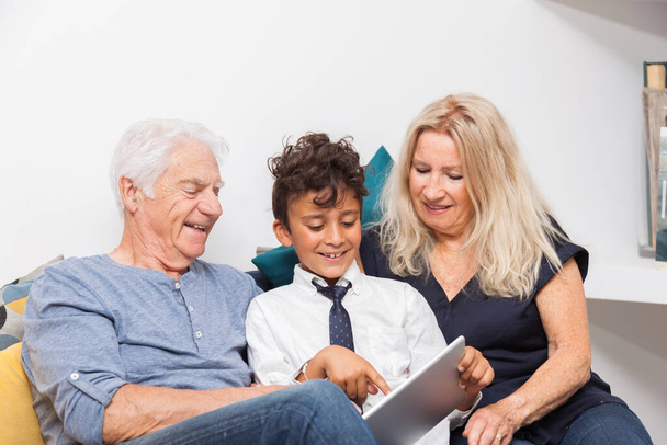 Autentyczny moment, kiedy chłopiec z babcią i jego dziadkiem bawią się cyfrowym tabletem na kanapie. Uśmiechnięta rodzina z tabletem w domu - Zdjęcie, obraz