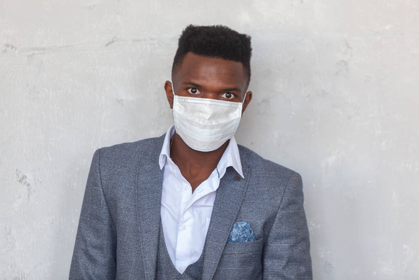 COVID-19. Confident young African man is wearing a protective medical face mask to prevent infection with coronavirus and pandemic standing in a grey suit against a concrete wall - Valokuva, kuva