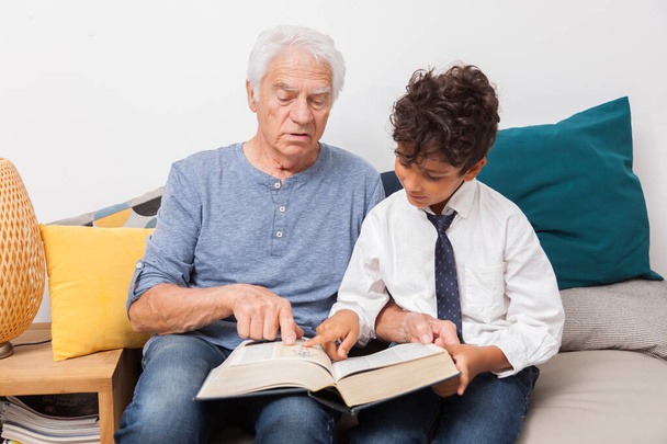 Szczęśliwy dziadek z wnukiem czytający razem książkę, literaturę na sofie. Koncepcja rodziny, kultury i radości - Zdjęcie, obraz