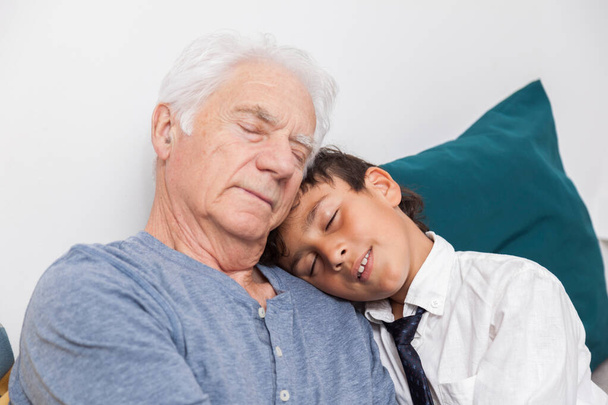 Happy boy cuddles his grandfather on a sofa. Concept of tenderness, family and joy - Photo, Image