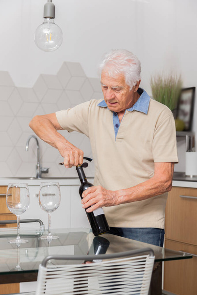 Senior öffnet Rotweinflasche mit Korkenzieher in Küche. - Foto, Bild
