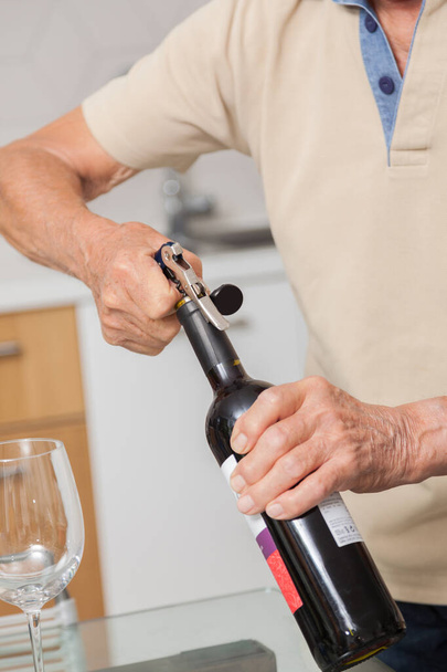 Senior otwierający czerwoną butelkę wina korkociągiem w kuchni. - Zdjęcie, obraz