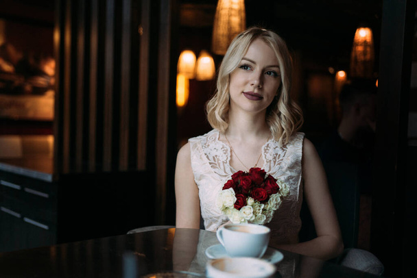 Beautiful young blonde women in wedding dress with rose bouquet in the cafe looking in the window - Photo, Image