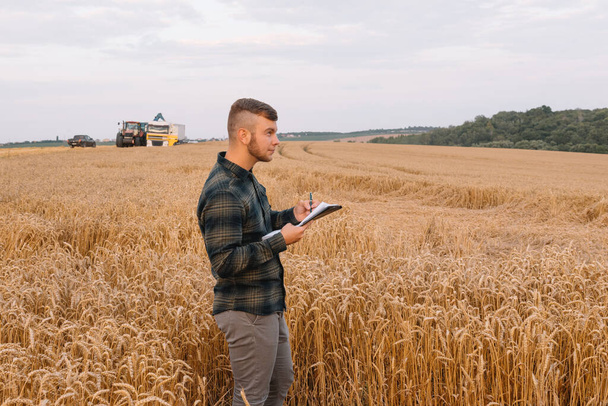 Young attractive farmer with laptop standing in wheat field with combine harvester in background - Foto, Imagem