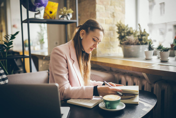 Woman freelancer leafing through pages of a diary in a coffee shop. Entrepreneur working in cafe. Business woman uses paper reminders and schedules. Girl writes notes diary, makes business plan. - Photo, Image
