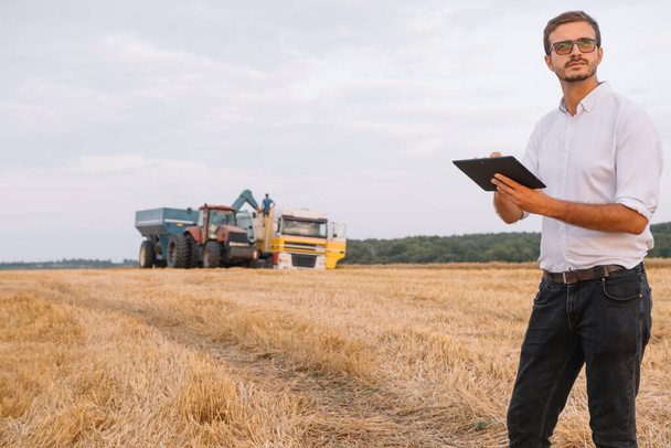 Szczęśliwy młody inżynier rolnik z notebooka stojącego na polu pszenicy podczas łączenia kombajn pracy w tle - Zdjęcie, obraz