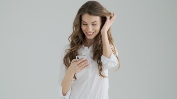 A positive beautiful young woman is using her smartphone isolated over a gray background in studio - Záběry, video