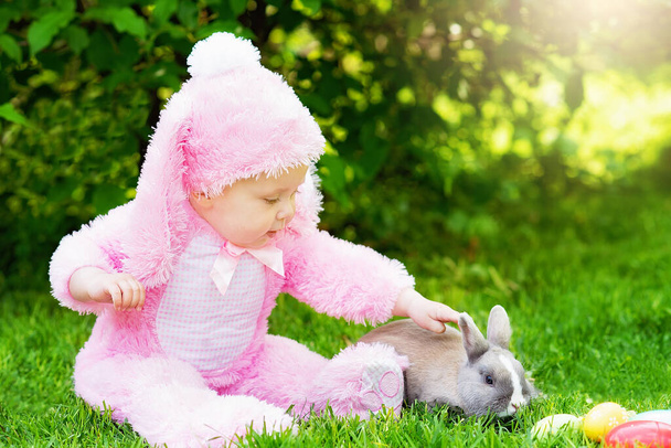  Kleines Mädchen im Hasenkostüm, das mit Hasen spielt, während es mit bemalten Ostereiern auf Gras im Garten sitzt. Urlaubskonzept  - Foto, Bild