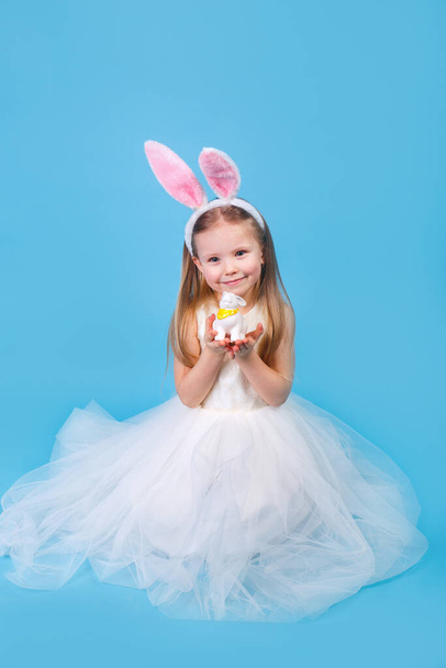 Cute positive little girl wearing bunny ears holding easter rabbit on blue studio background. Happy Easter - Photo, Image