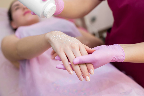 Close-up de um esteticista polvilha talco em uma mão de meninas jovens antes do procedimento de depilação. A menina está em um sofá em um salão de beleza, ela faz o procedimento shugaring
. - Foto, Imagem