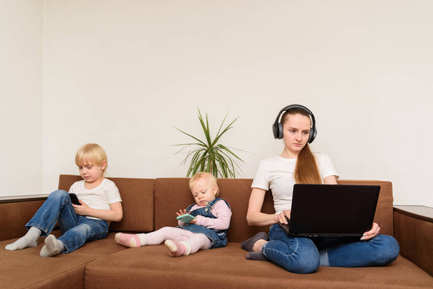 Jeune mère avec ordinateur à la main et les enfants avec des téléphones assis sur le canapé. Surutilisation des dispositifs
. - Photo, image
