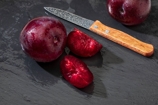 Appetizing and healthy red plums, whole and cut into segments with a rustic appearance on a black background. - Фото, изображение