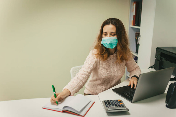 Frau in Schutzmaske, sitzt an einem Tisch mit Laptop und Taschenrechner und schützt Büroangestellte vor Viren, während der Quarantäne, das Konzept der Gesundheit und Coronavirus covid-19 - Foto, Bild