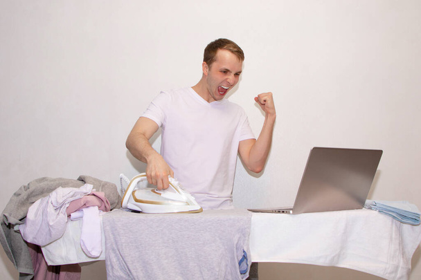 ένας νεαρός άνδρας στο σπίτι σε αυτο-απομόνωση εργάζεται εξ αποστάσεως και ασχολείται με το σιδέρωμα. - Φωτογραφία, εικόνα