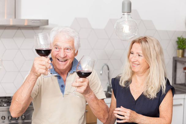 Alegre hombre mayor sosteniendo dos copas de vino tinto y su esposa, una hermosa mujer rubia se ríe en la cocina
 - Foto, Imagen