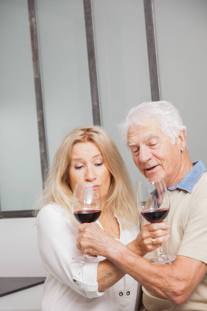 Happy senior couple relax talking and drinking red wine glasses together in the kitchen at home - Foto, Imagem