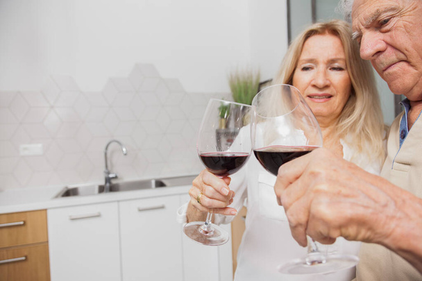 Feliz pareja de ancianos relajarse hablando y bebiendo copas de vino tinto juntos en la cocina en casa
 - Foto, Imagen