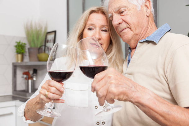 gelukkig senior paar ontspannen praten en het drinken van rode wijn glazen samen in de keuken thuis - Foto, afbeelding