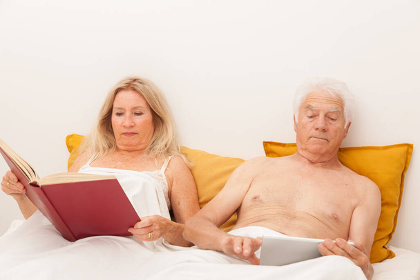 Yaşlı çift yatak odasında kitap okuyor ve tablete bakıyor. - Fotoğraf, Görsel