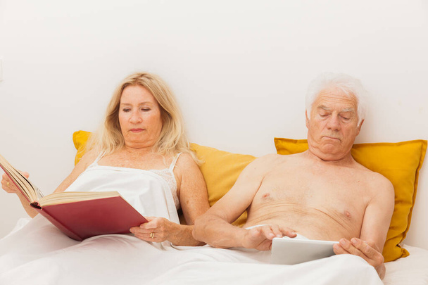 Старша пара читає і дивиться на планшет в ліжку в спальні
 - Фото, зображення