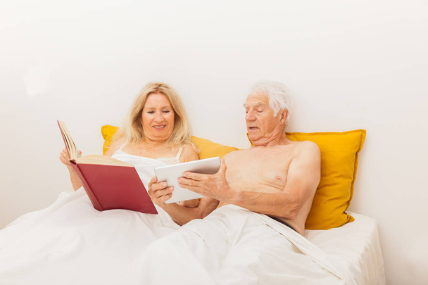 Ηλικιωμένο ζευγάρι διαβάζει και ψάχνει σε ένα tablet στο κρεβάτι σε ένα υπνοδωμάτιο - Φωτογραφία, εικόνα