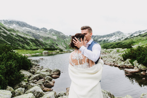 Mooie bruid en bruidegom knuffel en kus in de bergen in de buurt van het meer. - Foto, afbeelding