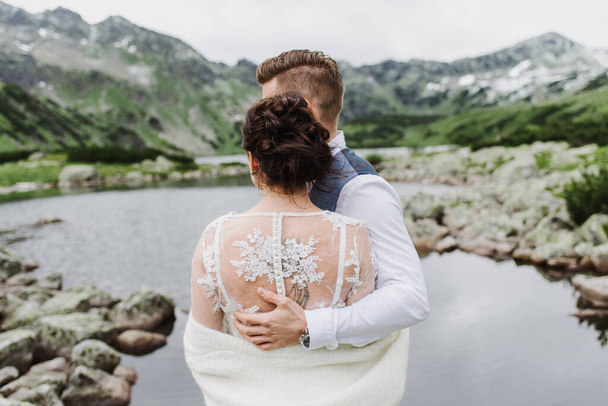 Όμορφη νύφη και γαμπρός αγκαλιά και φιλί στα βουνά κοντά στη λίμνη. - Φωτογραφία, εικόνα