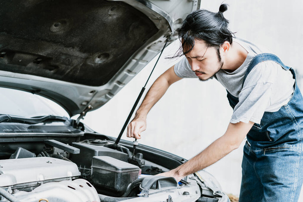 Young Asian handsome mechanic in uniform fixing a broken engine at the garage. Repair of machines, fault diagnosis, repair specialist,technical maintenance and people concept. - Φωτογραφία, εικόνα
