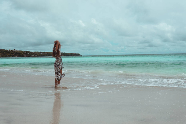 Mooi en jong meisje in een jurk poseren op de oever van de oceaan en de zee met blauwe golven. Vrouw in een lange jurk model en mode poseren. Ontspan en ontspan op Bali - Foto, afbeelding
