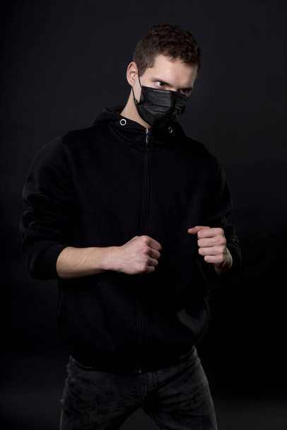 Man in black in a fighting pose with clenched fists. On the young mans face, a black medical mask covers his nose and mouth. - Fotoğraf, Görsel