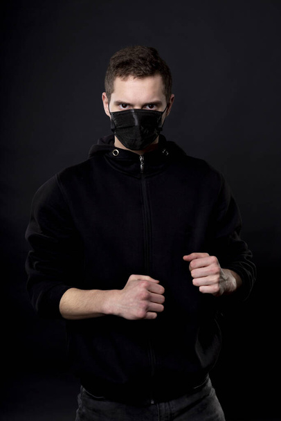 Man in black in a fighting pose with clenched fists. On the young mans face, a black medical mask covers his nose and mouth. - Photo, Image