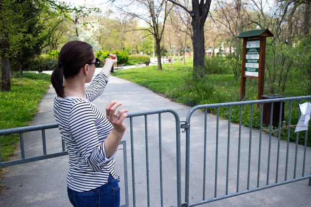  Νεαρή γυναίκα μπροστά από ένα κιγκλίδωμα και μια πινακίδα που απαγορεύει την είσοδο στο πάρκο. Παιδικές χαρές και δημόσια πάρκα έκλεισαν λόγω πανδημίας της νόσου του Coronavirus COVID-19. Δεν επιτρέπεται η είσοδος. Σερβία. - Φωτογραφία, εικόνα