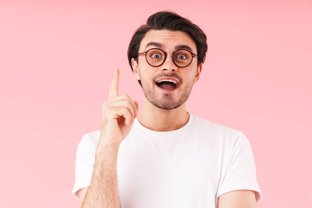 Image of brunette amazed man wearing eyeglasses pointing finger upward at copyspace isolated over pink background - 写真・画像