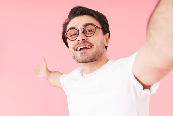 Image of young happy man wearing eyeglasses smiling and taking selfie photo isolated over pink background - Фото, зображення