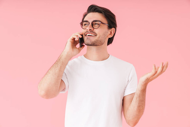 Image of young pleased man wearing eyeglasses talking on cellphone and smiling isolated over pink background - Foto, Imagen
