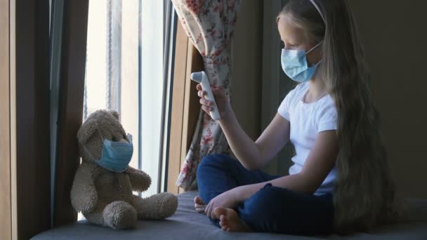 Stay at home quarantine coronavirus pandemic prevention. Sad girl and his teddy bear both in protective medical masks sitting near the window, a girl measures the temperature of the bear. - Video
