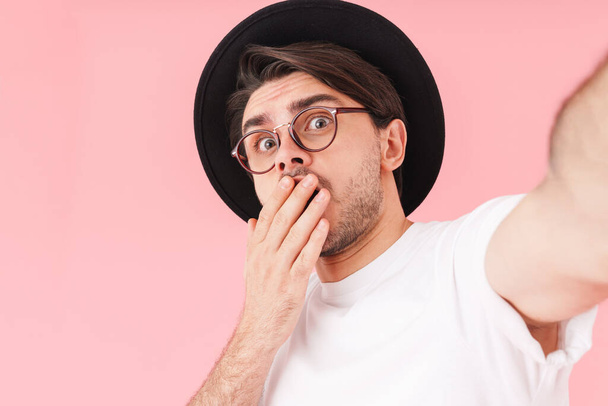 Image of shocked man wearing eyeglasses and hat expressing fear while taking selfie photo isolated over pink background - Zdjęcie, obraz