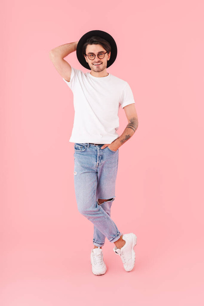 Image of young cheerful man wearing eyeglasses and black hat poising and smiling at camera isolated over pink background - Fotografie, Obrázek