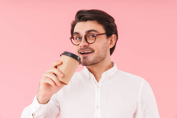Image of caucasian joyful man wearing eyeglasses drinking coffee from paper cup and smiling isolated over pink background - Foto, imagen