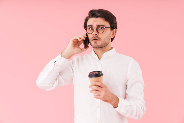 Image of caucasian upset man wearing eyeglasses drinking coffee takeaway and talking on cellphone isolated over pink background - Fotografie, Obrázek