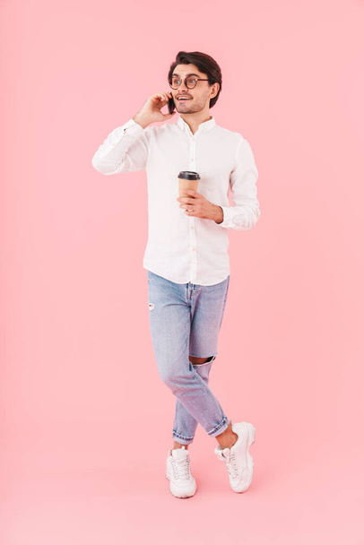 Image of unshaven happy man wearing eyeglasses drinking coffee takeaway and talking on cellphone isolated over pink background - Photo, Image