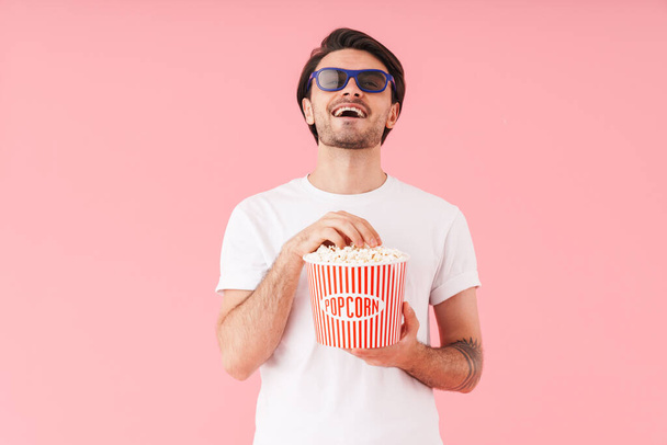 Image of young laughing man wearing 3d glasses eating popcorn while watching movie isolated over pink background - 写真・画像