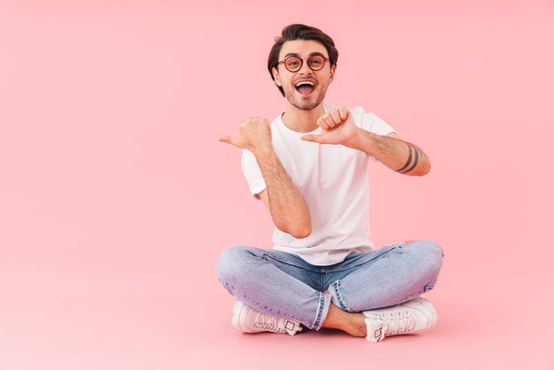 Image of joyful man wearing eyeglasses pointing fingers at copyspace while sitting with legs crossed isolated over pink background - Foto, Imagem