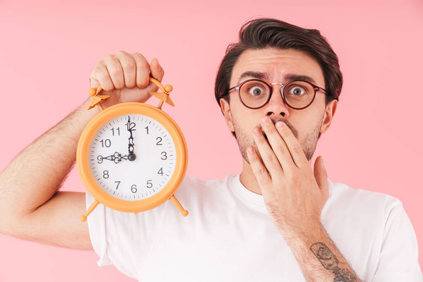 Image of stressed unhappy man wearing eyeglasses holding desk alarm and looking at camera isolated over pink background - Foto, Imagem