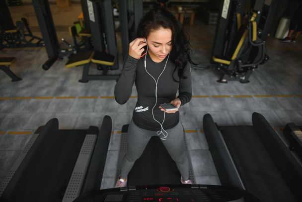 Young fit woman running on treadmill listening to music via headphone at gym. Concept of healthy lifestyle. - Foto, afbeelding